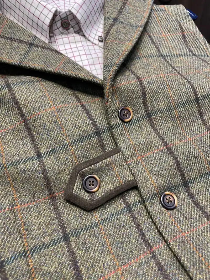 Close-up detail of custom green tweed shooting waistcoat showing windowpane check pattern, horn buttons, hexagonal cartridge pocket with contrasting trim, and notched lapel collar