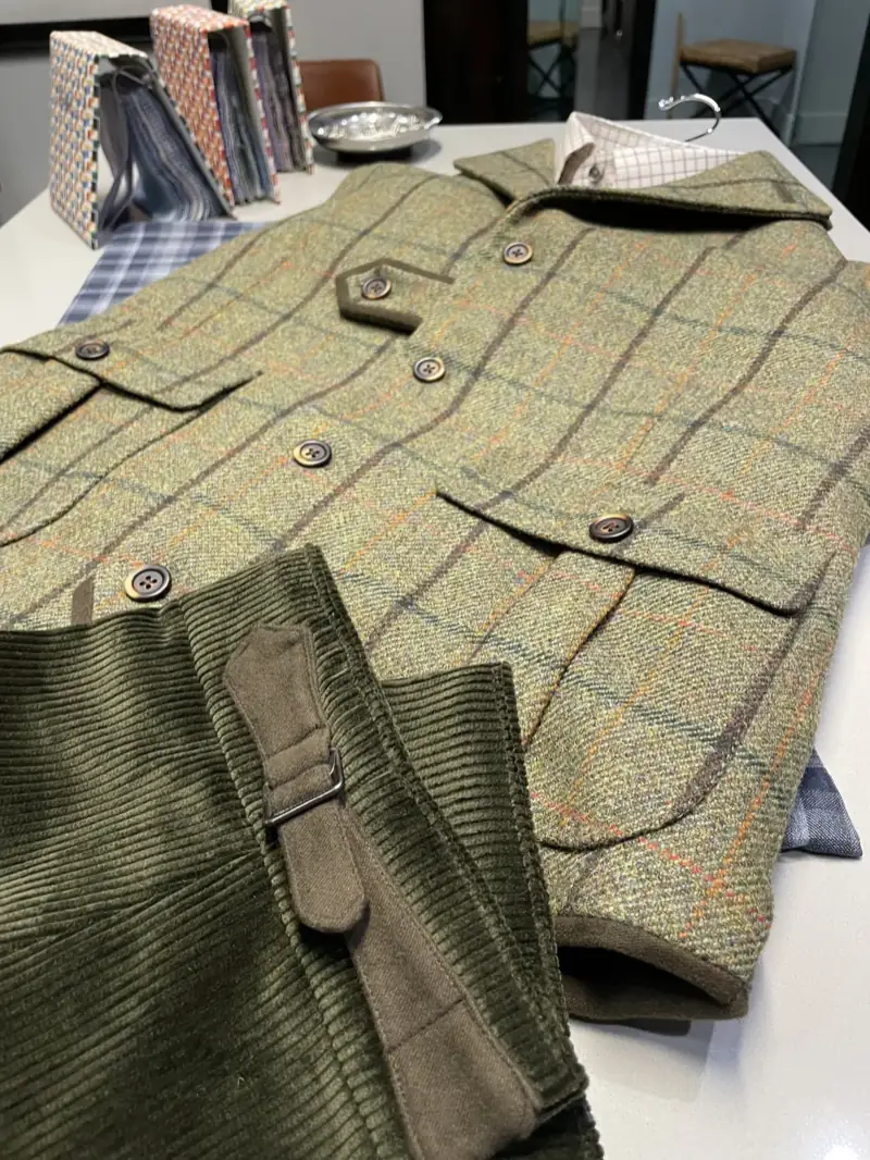 Flat-lay of custom olive green tweed shooting vest with five-button front, hexagonal cartridge pocket, flapped bellows pockets, and horn buttons, paired with olive corduroy trousers on tailor's cutting table